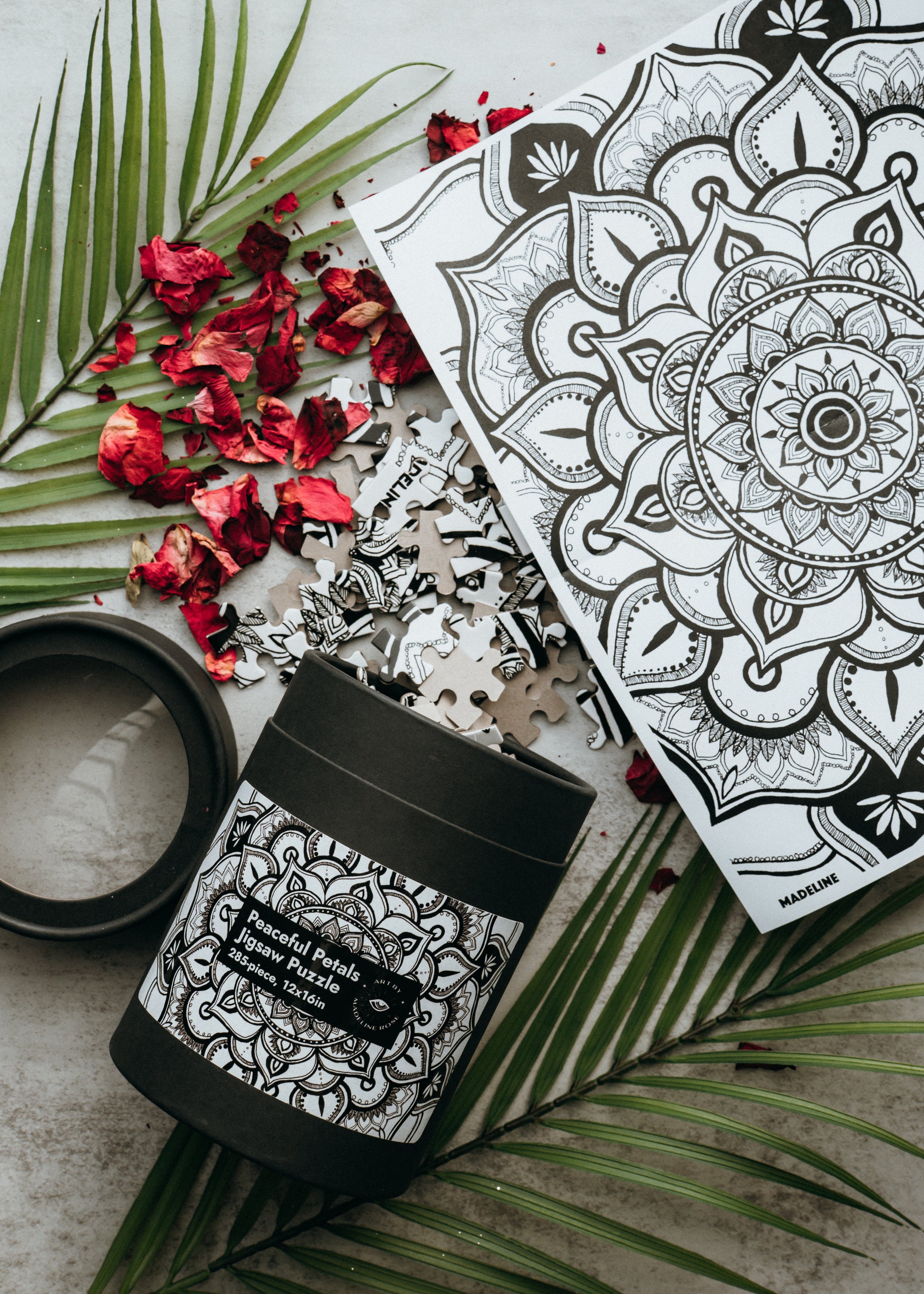 A jigsaw puzzle with interlocking pieces spread out, forming a floral pattern mandala. The puzzle is packaged in a black kraft tube with a label showing the finished image.