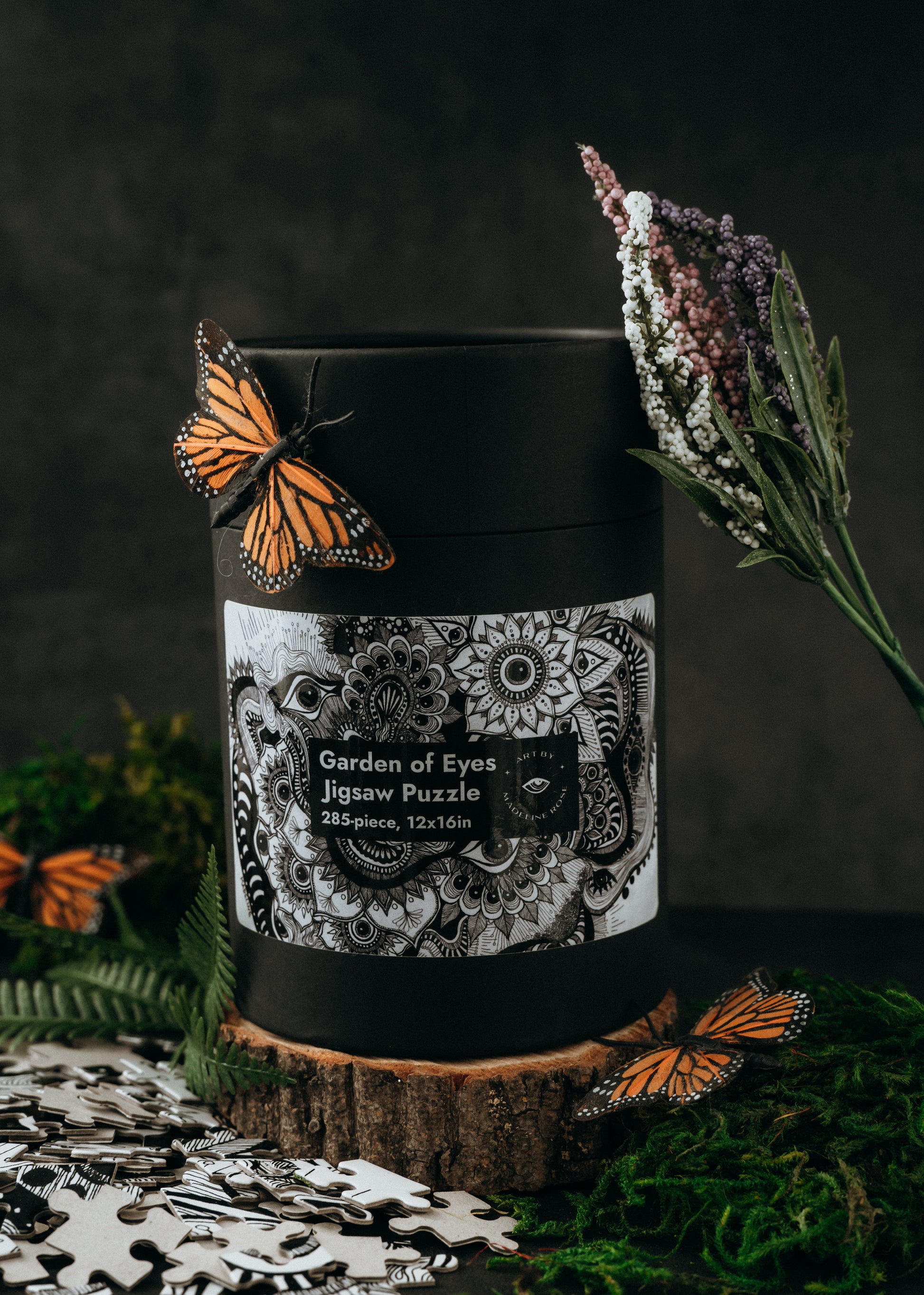 A completed 285-piece jigsaw puzzle depicting a Garden of Eyes theme with butterflies and floral elements, presented in a black cylindrical container with a label and a butterfly resting on it.
