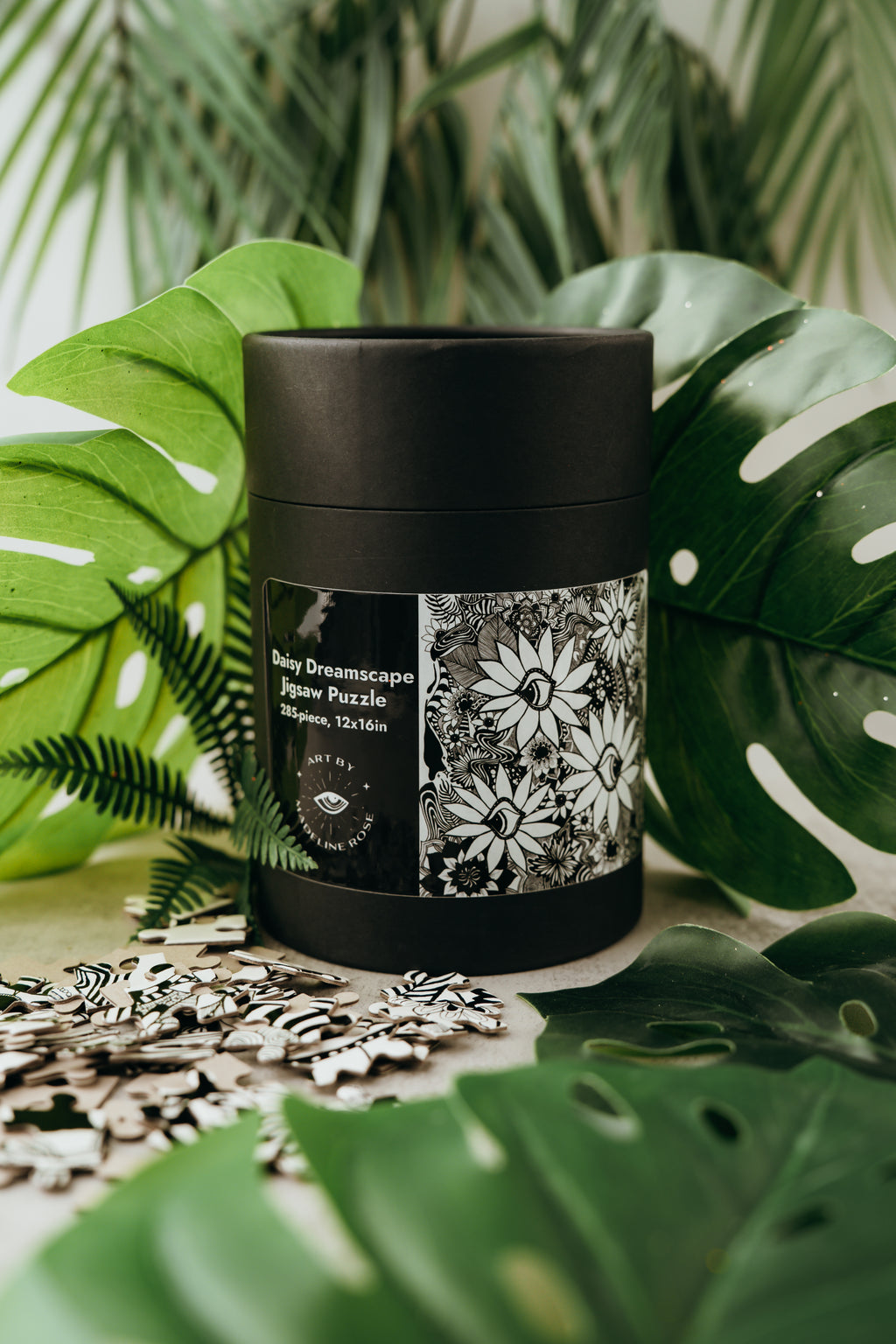 A 285-piece jigsaw puzzle depicting a floral pattern, specifically daisies, in a black cylindrical container with a label that reads 'Daisy Dreamscape'. The container is placed on a table with green leaves in the background.